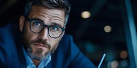 Thoughtful businessman with glasses and gray beard working late at office, holding pen while contemplating important decision in dark environment.