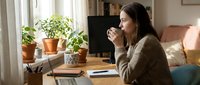 Woman with dark hair and medium complexion, wearing a beige cardigan, sitting at wooden desk with laptop, drinking coffee while working from home office with potted plants and window light.