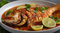 Grilled whole fish with spicy tomato sauce, lime wedges and fresh herbs in white bowl. Traditional seafood dish ready for restaurant menu or cooking blog.