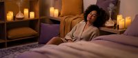 Woman with curly dark hair, medium brown skin tone, wearing beige cardigan, sitting on bed surrounded by lit candles in warm bedroom setting for relaxation and wellness content.