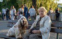 Senior woman sitting on park bench with cocker spaniel dog, enjoying peaceful afternoon in busy public garden.
