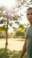 Middle-aged man of Latin American ethnicity wearing casual gray t-shirt standing in sunlit park with trees and bokeh background, outdoor portrait for lifestyle and wellness concepts.