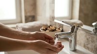 Young adult with medium skin tone washing hands under running water at bathroom sink with chrome faucet, promoting hygiene and cleanliness routines for health and wellness campaigns.