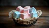 Soft pastel marshmallows in pink and blue colors arranged in rustic wire basket with parchment paper on wooden surface, creating cozy sweet atmosphere.