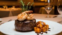 Grilled steak topped with shrimp and rosemary garnish, served with roasted vegetables on elegant white plate in fine dining restaurant setting.