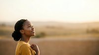 Young woman of African descent wearing yellow shirt gazing upward at sunset over rural field, expressing hope, contemplation, and peaceful solitude in nature.