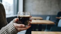 Person holding glass mug of dark coffee in cozy cafe setting with warm sweater sleeves visible and blurred background tables for relaxation concept.