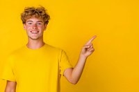 Young Caucasian male with curly blonde hair wearing yellow t-shirt points finger to side while smiling against vibrant yellow background. Modern minimal studio portrait.