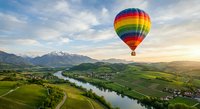 Colorful hot air balloon floating over scenic valley with winding river, green fields and mountain ranges during golden hour sunset creating peaceful landscape.
