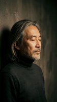 Mature Asian man with gray hair and beard in contemplative pose wearing dark turtleneck sweater against textured wall background for lifestyle photography.