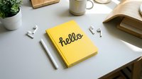 Yellow notebook with hello lettering on a clean modern desk with wireless earbuds, pen, plant, and wooden accessories in a minimal workspace.