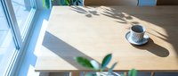 Wooden desk with coffee cup and plant near bright window, natural sunlight creating shadows, minimalist workspace setup for office or home environment.
