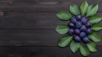 Fresh ripe plums arranged with green leaves on dark wooden background, top view composition for food photography and menu design.