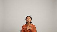 Young woman with dark hair and South Asian ethnicity wearing a rust-orange long-sleeve shirt, smiling with hands placed on chest in a grateful or appreciative gesture against neutral background.