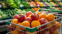 Grocery shopping cart filled with fresh red apples and oranges at supermarket produce section with blurred shelves background.