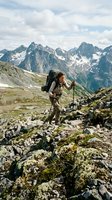 Hiker with backpack and trekking poles navigating rocky alpine terrain with dramatic mountain peaks and snow patches in background, suitable for travel and outdoor adventure content.