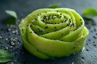 Delicate rose-shaped avocado garnish sprinkled with black sesame seeds on dark slate background, fresh green leaves accent culinary presentation.