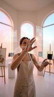 Female artist with paint-covered hands working on transparent glass surface in bright studio with arched windows and easels in background.