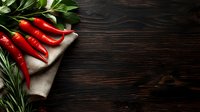 Fresh red chili peppers with green herbs on rustic linen napkin against dark wooden background. Natural food photography with copy space for text.
