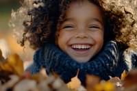 Joyful African American child with curly hair wearing blue knit scarf laughs heartily among autumn leaves, capturing pure happiness in warm natural lighting.