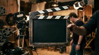 Filmmaker holding vintage film clapperboard on movie set with professional camera equipment and warm studio lighting in background for cinema production.