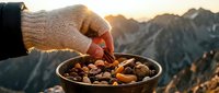 Person in warm sweater holding bowl of mixed nuts and dried fruits at mountain summit during golden hour sunset.