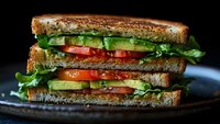 Fresh layered vegetable sandwich with toasted whole grain bread, ripe tomatoes, crisp lettuce, and sliced avocado on dark rustic plate against black background.