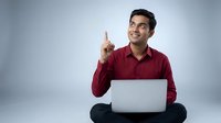 Young Indian businessman in red shirt pointing up with laptop, sitting cross-legged on floor against gray background. Business concept for presentations.