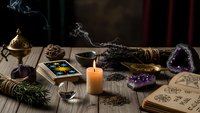 Mystical fortune telling setup with tarot cards, burning candle, amethyst crystals, sage bundle and ancient spell book on wooden table for divination.