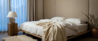 Modern minimalist bedroom interior with unmade bed, neutral beige tones, natural lighting from large window, wooden nightstand and contemporary home decor.