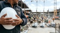Male construction worker holding white safety helmet at active building site with cranes and equipment, representing workplace safety and professional construction industry.