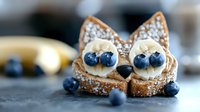 Creative breakfast toast shaped as fox face with banana slices, blueberries, and powdered sugar on dark background, fun food art for children.