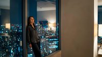 Professional businesswoman in black suit standing by floor-to-ceiling windows overlooking illuminated city skyline at night in modern office building.