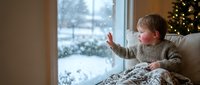 Young blonde boy in cozy sweater touching snowy window during winter holidays with Christmas tree lights creating warm festive atmosphere at home.