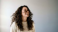 Young woman with curly hair laughing joyfully in cream sweater against neutral background for lifestyle and wellness content.