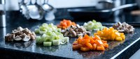 Fresh diced vegetables including carrots, celery, mushrooms and bell peppers arranged on dark cutting board in professional kitchen for cooking preparation.