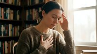 Young woman experiencing stress and anxiety sitting by window in cozy library setting with books in background for mental health awareness campaigns.
