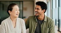 Young diverse couple having friendly conversation in modern office setting, man of Middle Eastern descent wearing olive shirt and woman of Asian descent in beige cardigan, both smiling during casual workplace interaction.