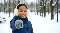 Happy African American boy in winter coat and earmuffs pointing at camera in snowy park, expressing joy and excitement during cold weather outdoor activity.