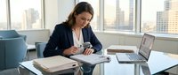 Professional businesswoman working at modern office desk with laptop and documents, reviewing paperwork in corporate environment with city skyline view.