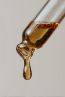 Golden amber liquid drop forming at tip of glass dropper against neutral background, capturing moment before falling in macro photography.
