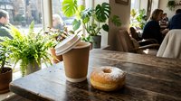 Takeaway coffee cup and fresh donut on wooden table by window in bright cafe with potted plants and blurred customers in background, suitable for food and beverage marketing.