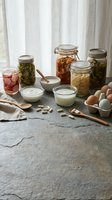 Fermented foods and probiotic supplements on stone surface, including kimchi, pickled vegetables, yogurt, sauerkraut in jars, eggs, and capsules.