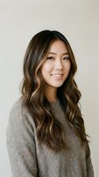 Young Asian woman with long wavy brown hair and highlights smiling warmly in cozy beige sweater against neutral background for lifestyle and portrait photography.