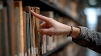 Female hand in gray sweater selecting book from library shelf, with soft bokeh background and warm lighting creating cozy atmosphere. Useful for education and literature concepts.