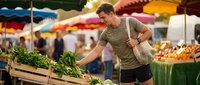Young Caucasian man shopping for fresh vegetables at outdoor farmers market with colorful umbrellas and produce stands in background.