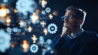 Young Caucasian businessman wearing glasses contemplates innovative technology solutions with glowing gears and data analytics interface floating in dark office space.