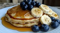 Stack of golden pancakes topped with fresh blueberries and sliced bananas, drizzled with maple syrup on gray ceramic plate, close up view of breakfast meal.