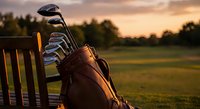 Golf bag with clubs on wooden bench at sunset course with warm golden light and green fairway background for sports equipment advertising.