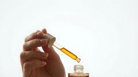 Hand holding glass dropper with golden oil serum over open bottle against white background for skincare and cosmetic product design.